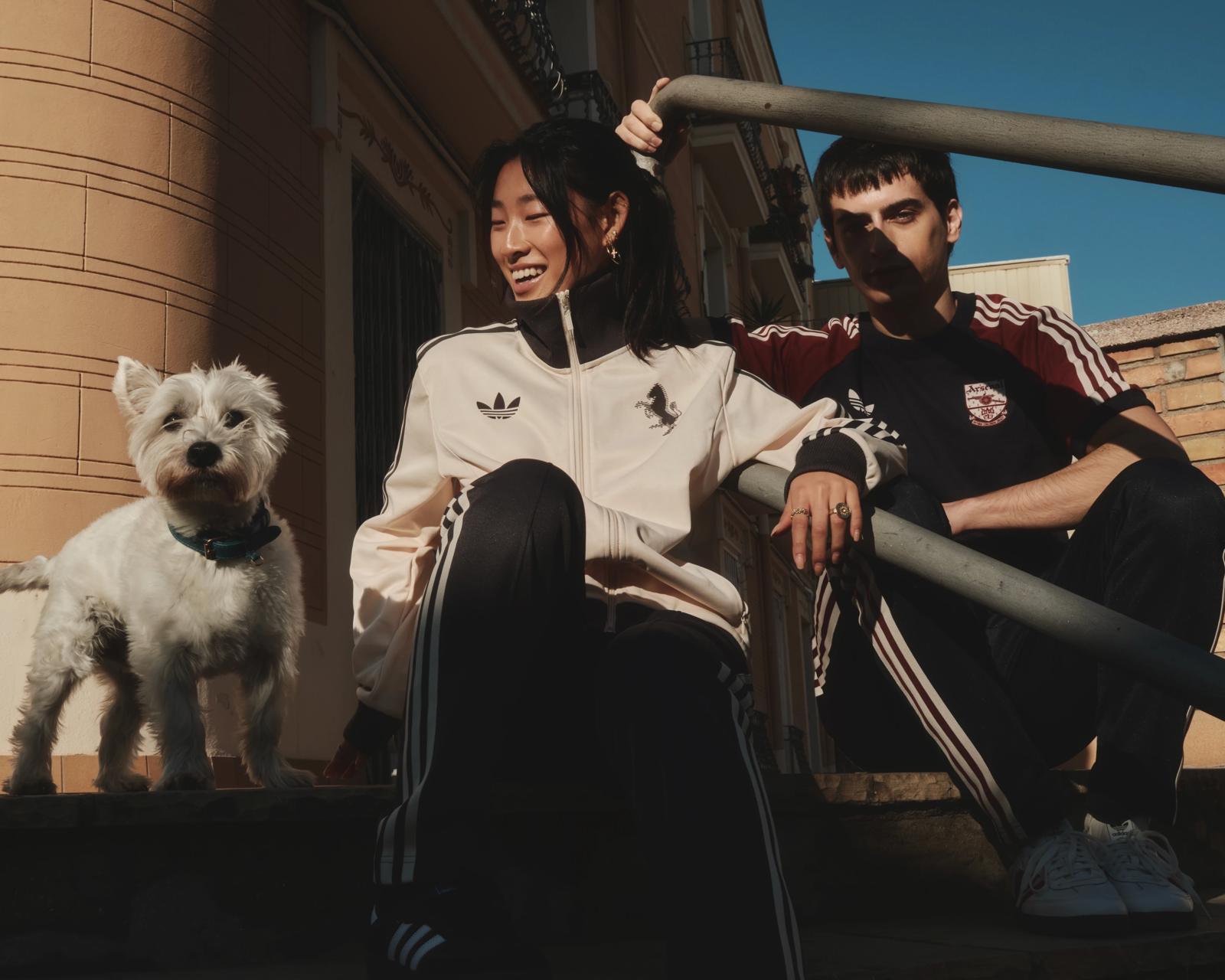 Dos modelos con chándales Adidas sentados en unas escaleras junto a un perro blanco