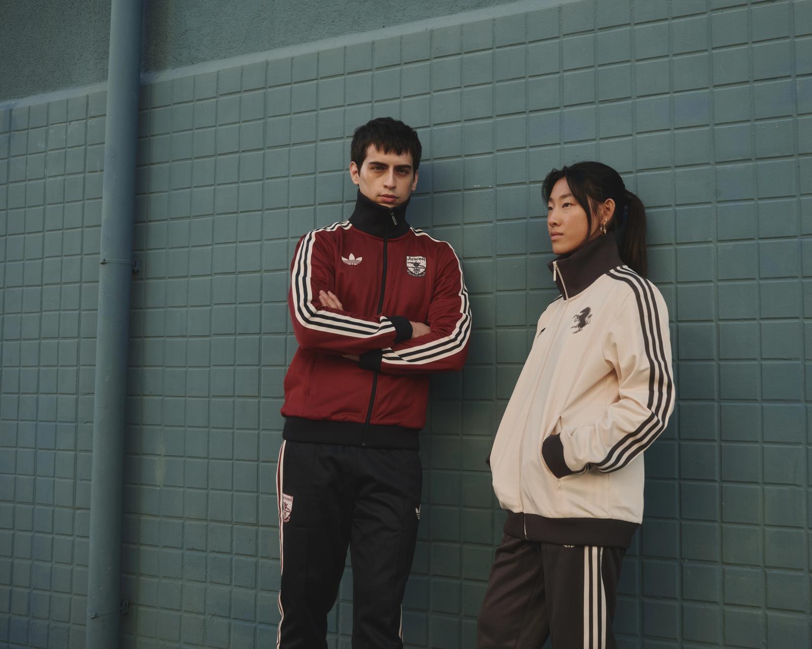 Dos modelos con chaquetas Adidas apoyados en una pared de azulejos verdes