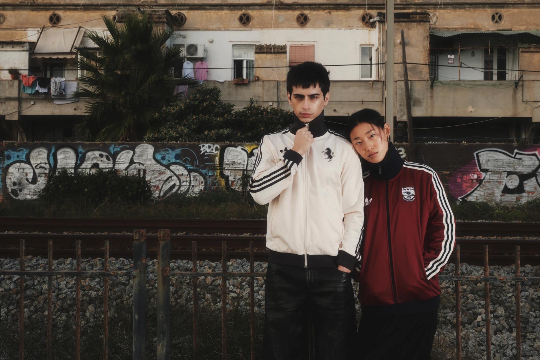 Dos modelos con chaquetas Adidas, uno apoyado en el hombro del otro, delante de un muro con grafitis