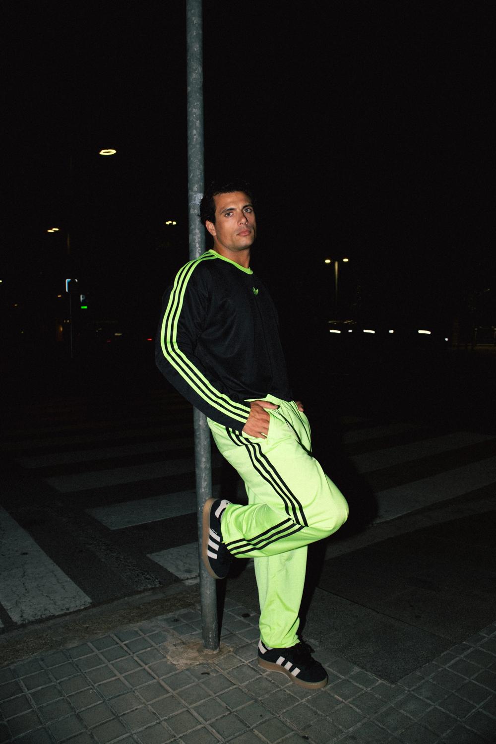 Modelo con pantalón Adidas amarillo flúor apoyado en un poste junto a un paso de cebra, de noche