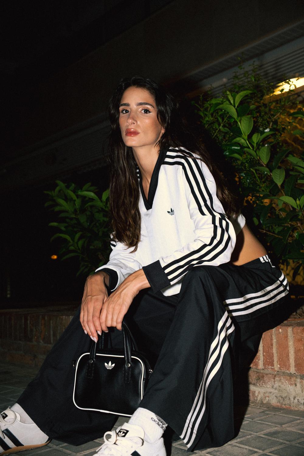 Modelo con camiseta Adidas blanca y pantalón negro sentada en un muro de ladrillo, con un bolso negro