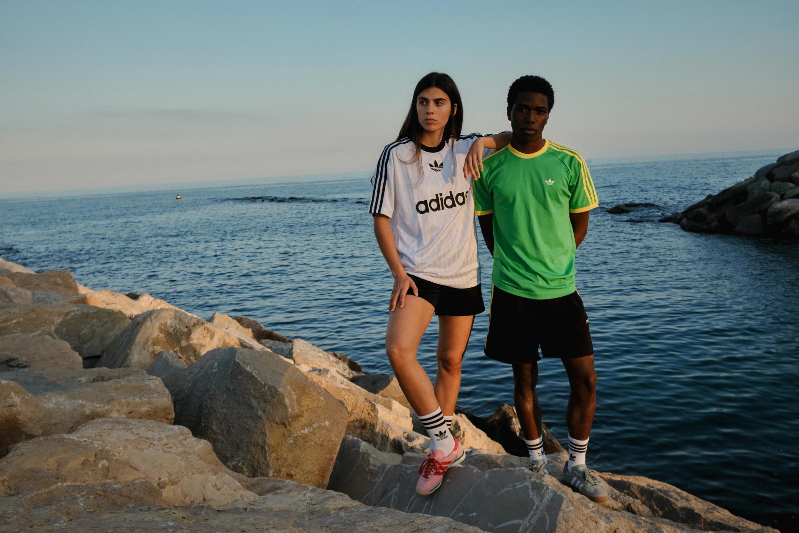 Dos modelos con camisetas Adidas de pie sobre un espigón de rocas, con el mar detrás