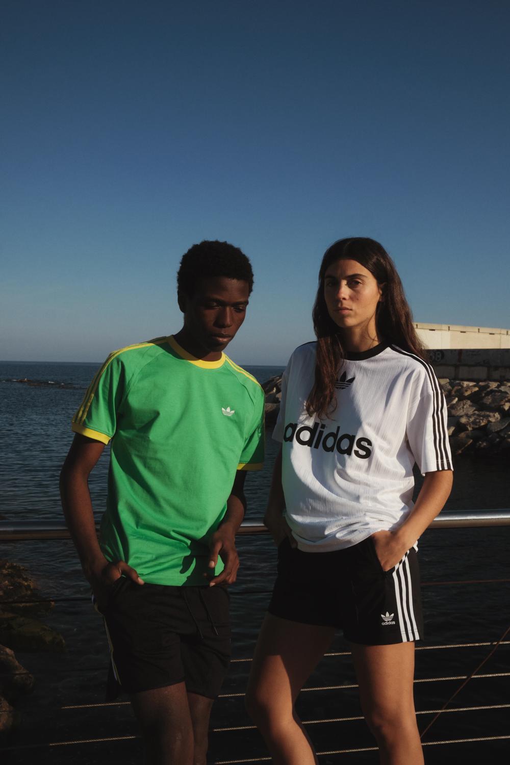 Dos modelos con camisetas Adidas apoyados en una barandilla frente al mar