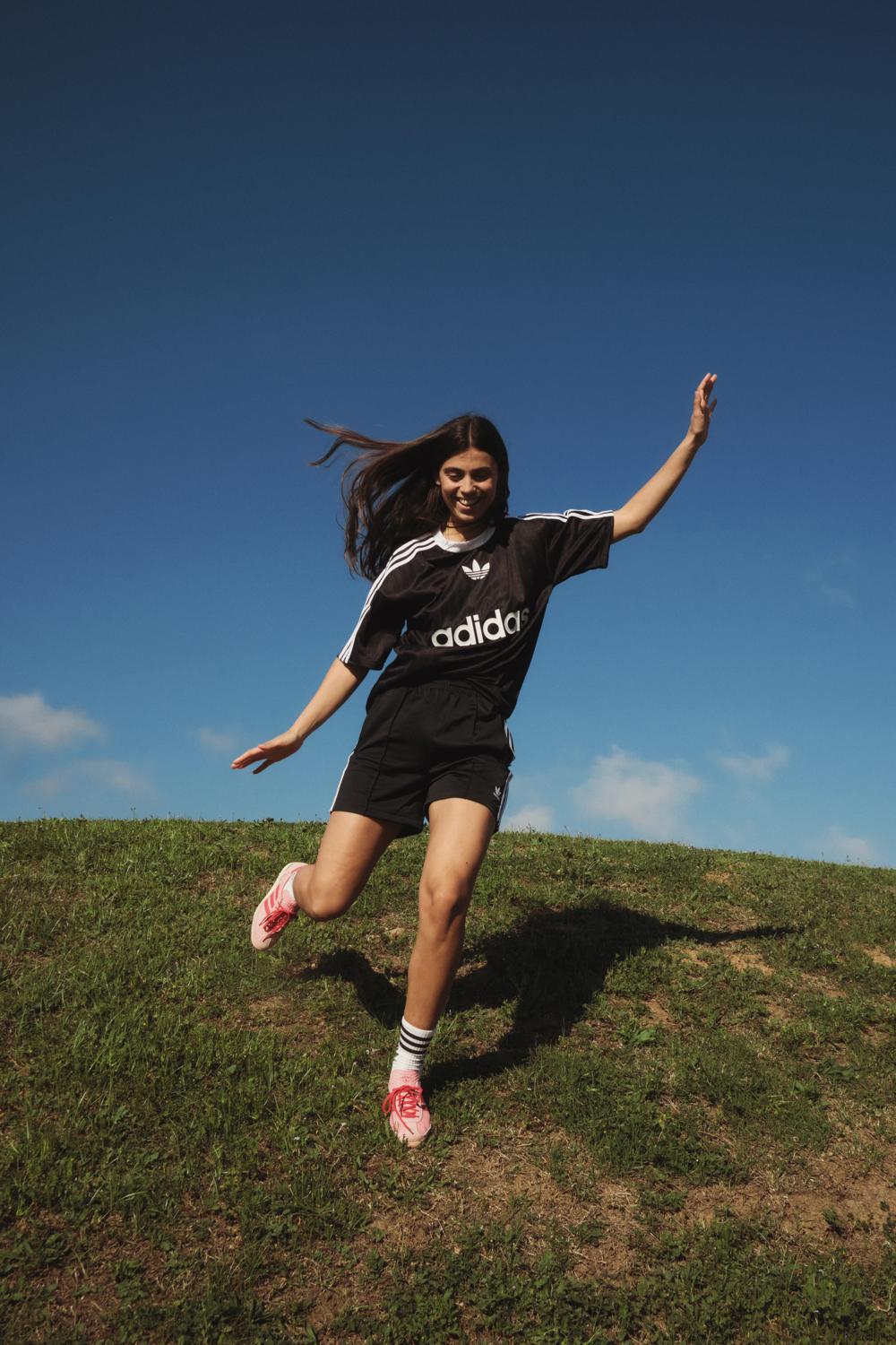 Modelo con equipación Adidas negra bajando una colina de césped bajo un cielo azul