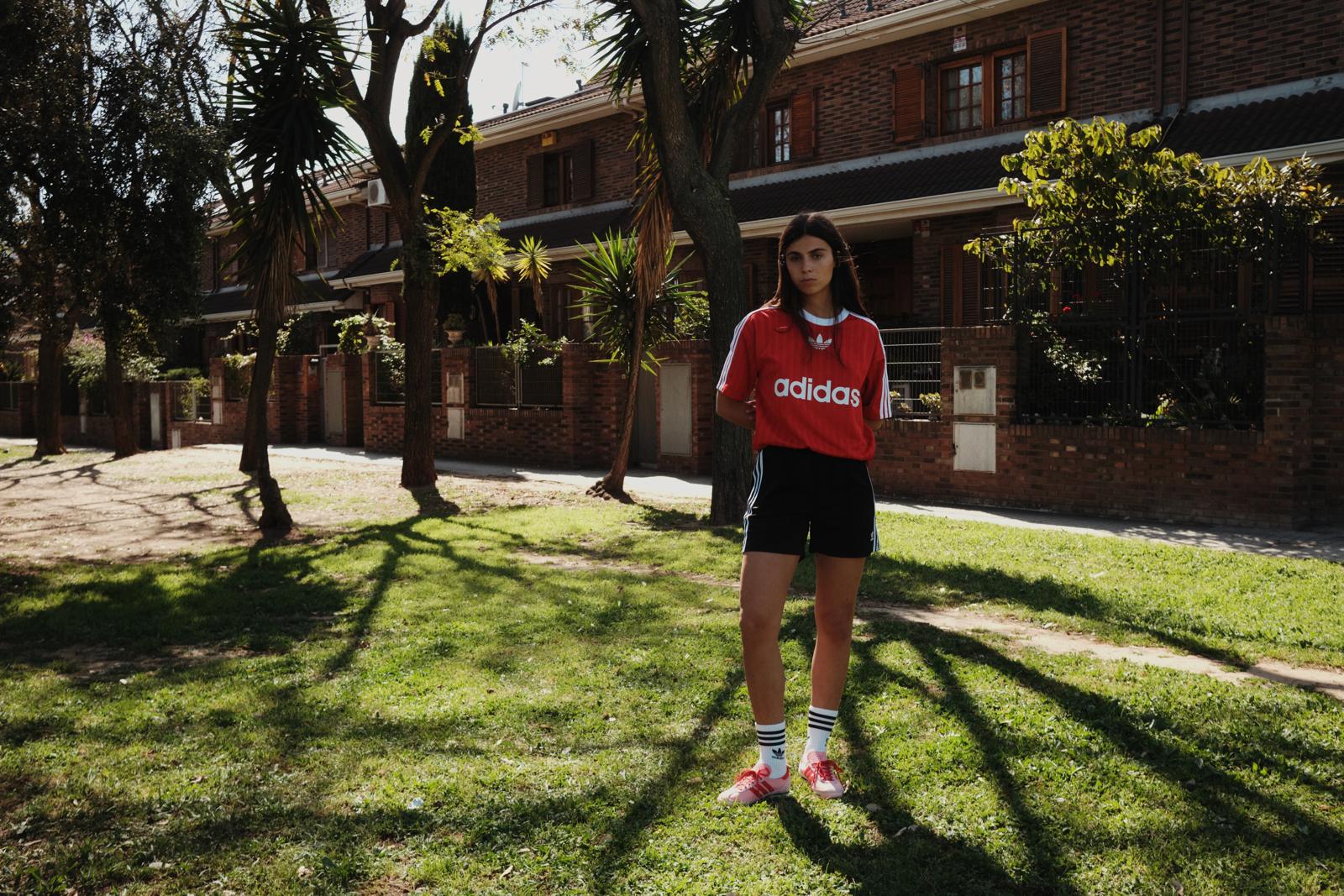 Modelo con camiseta Adidas roja en un jardín, delante de unas casas de ladrillo