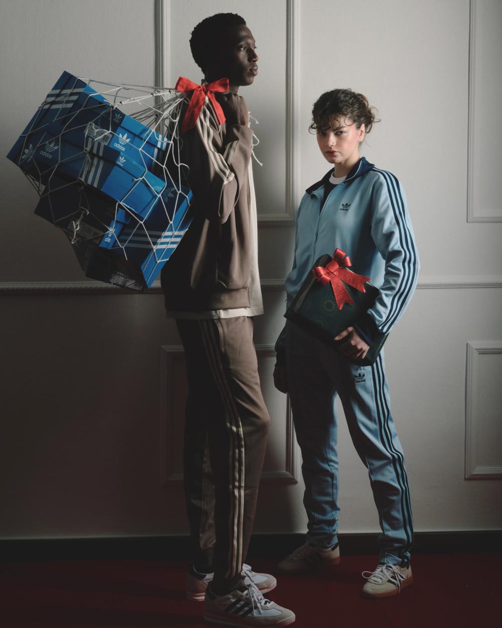 Dos modelos con chándales Adidas; uno carga una red con cajas de zapatillas y la otra sostiene un regalo