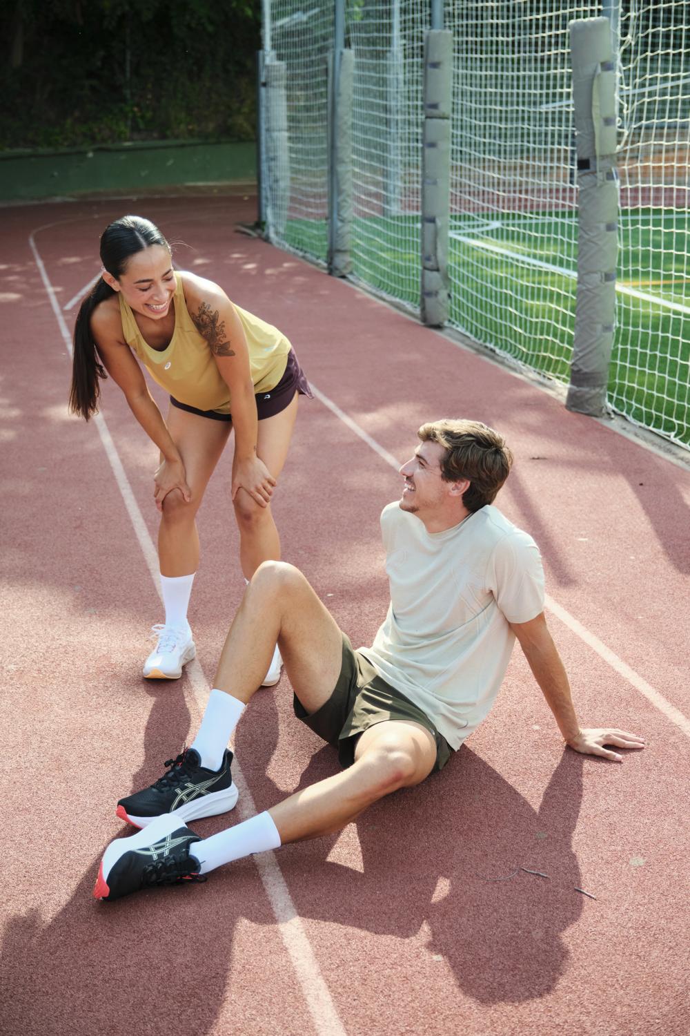 Una corredora de pie y un corredor sentado ríen en una pista de atletismo tras entrenar